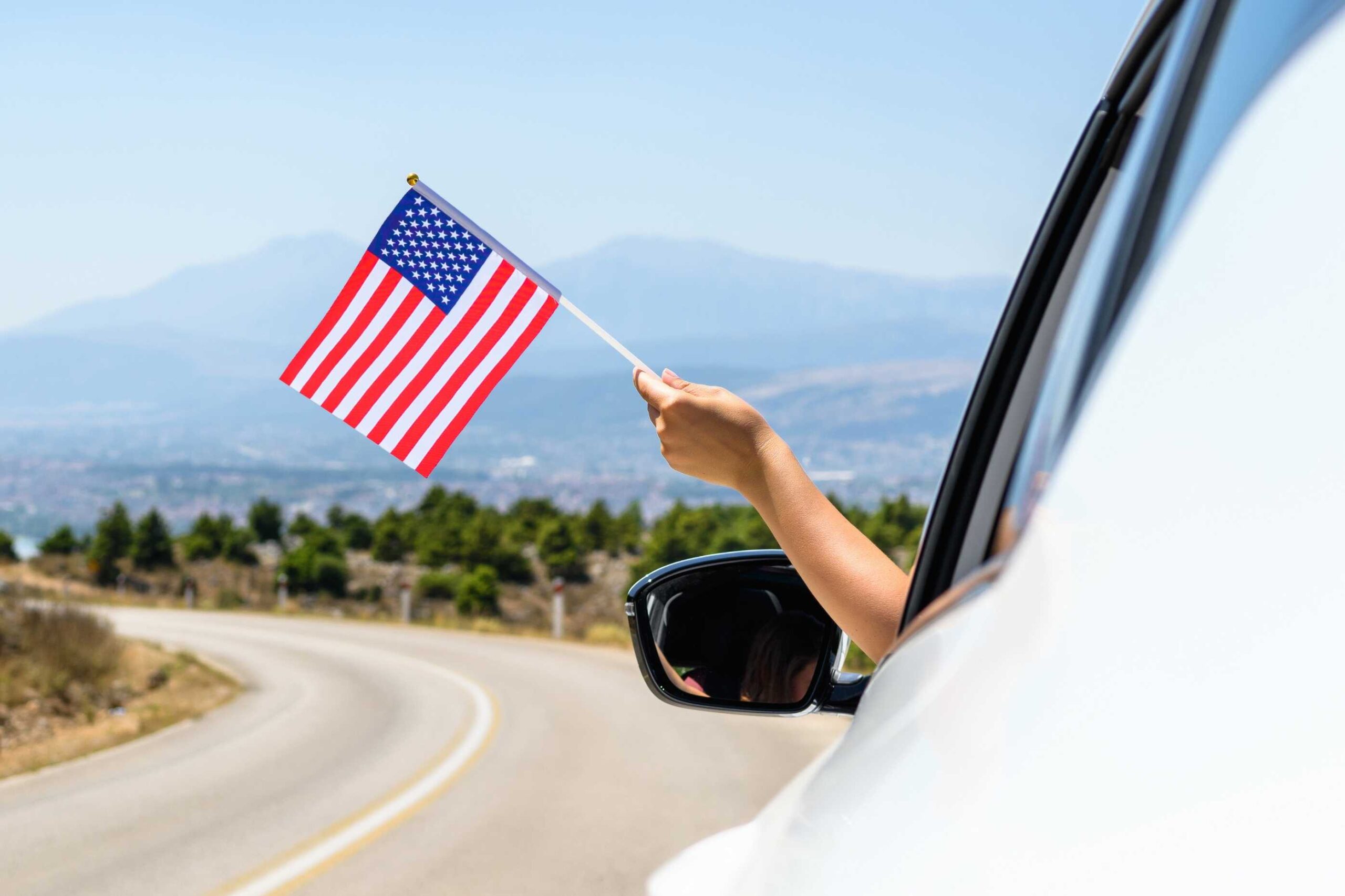 Person waving an American flag outside a car window with scenic road in the background, showing how important car rental excess insurance is for car renters.