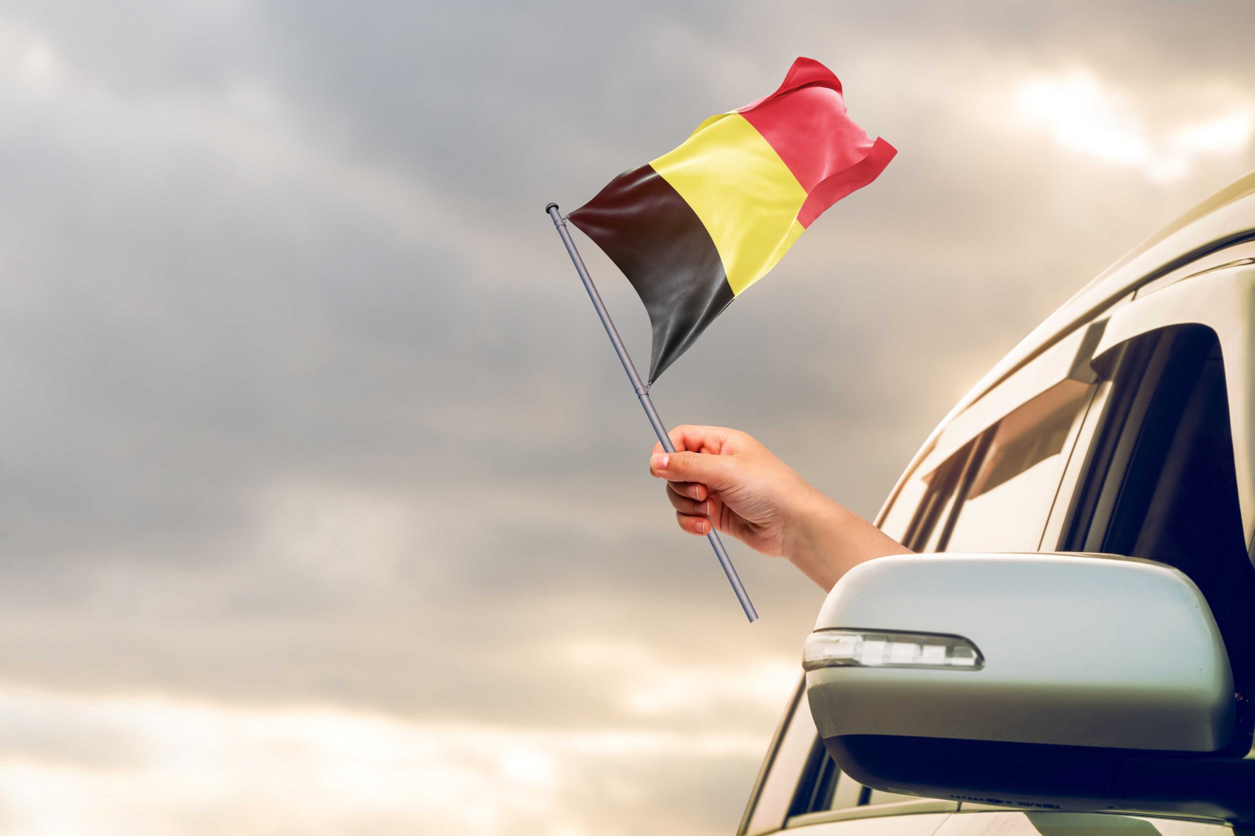 Person waving a Belgian flag from a car window with the cloudy sky behind, reinforcing the importance for people renting cars to get car rental excess insurance.