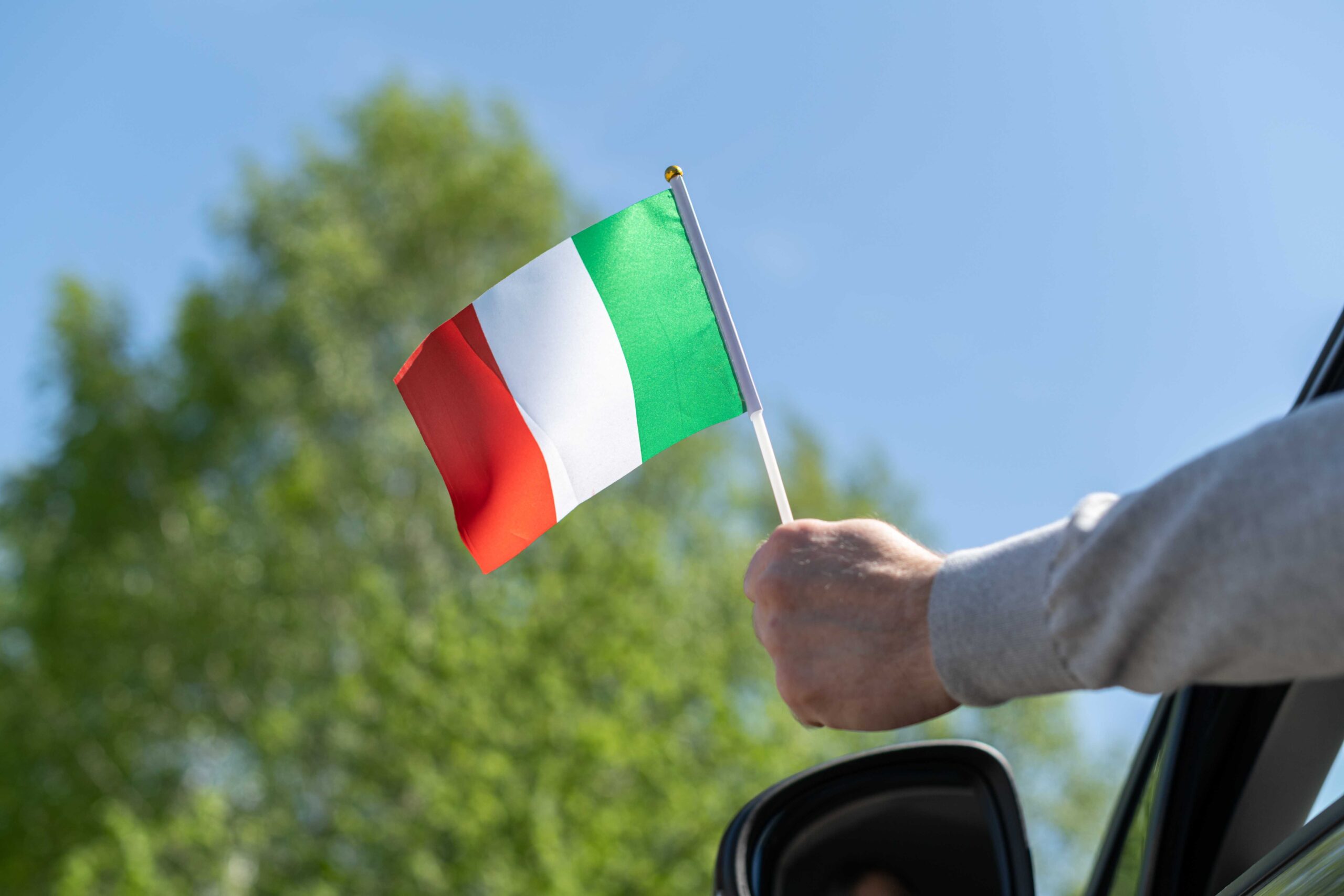 Person waving Italian flag out of car window with blue sky and green tree behind, emphasising importance of car rental excess insurance.