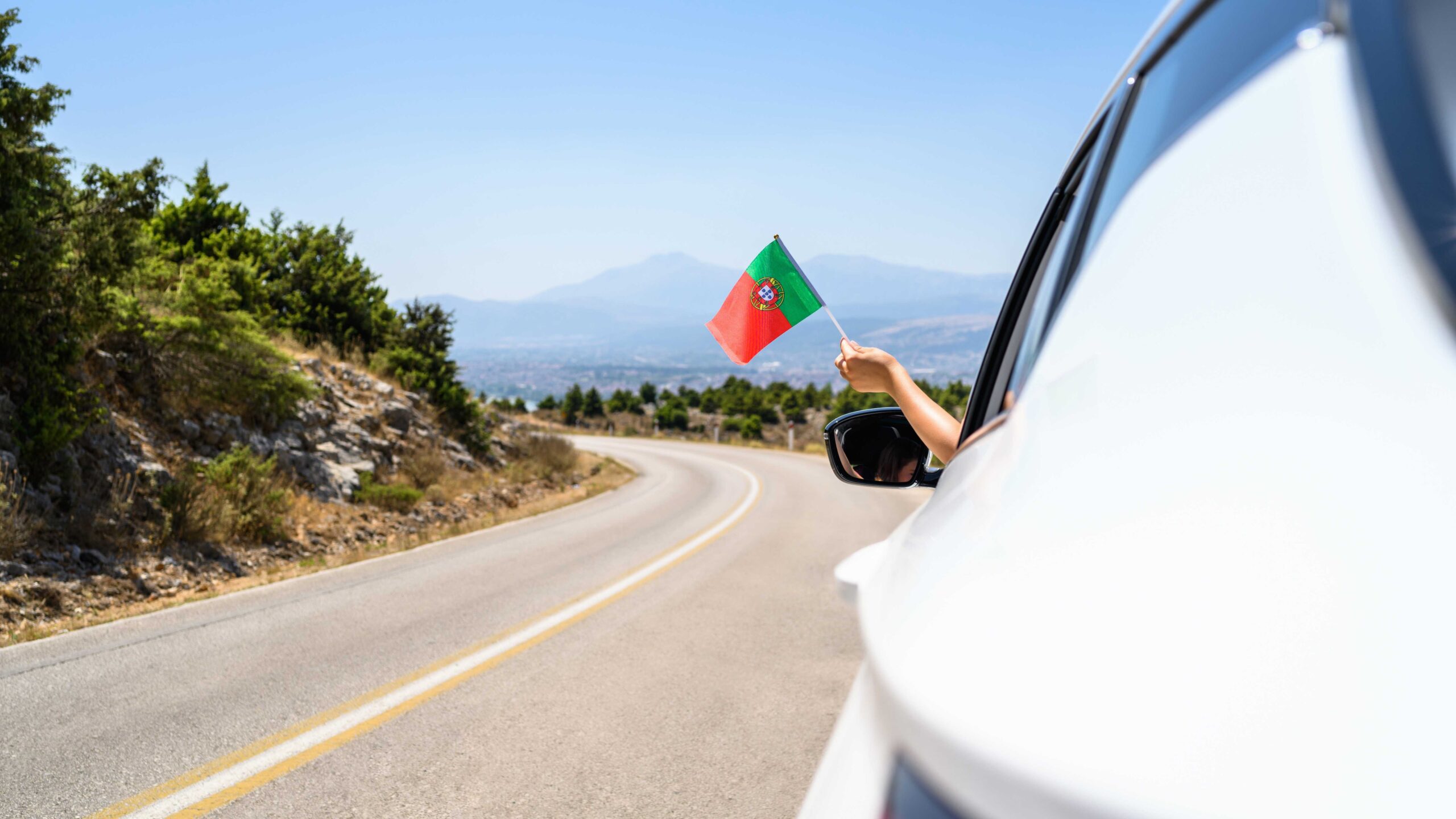 Person waving Portuguese flag outside a car window on scenic road underneath a blue sky, highlighting importance of car rental excess insurance.