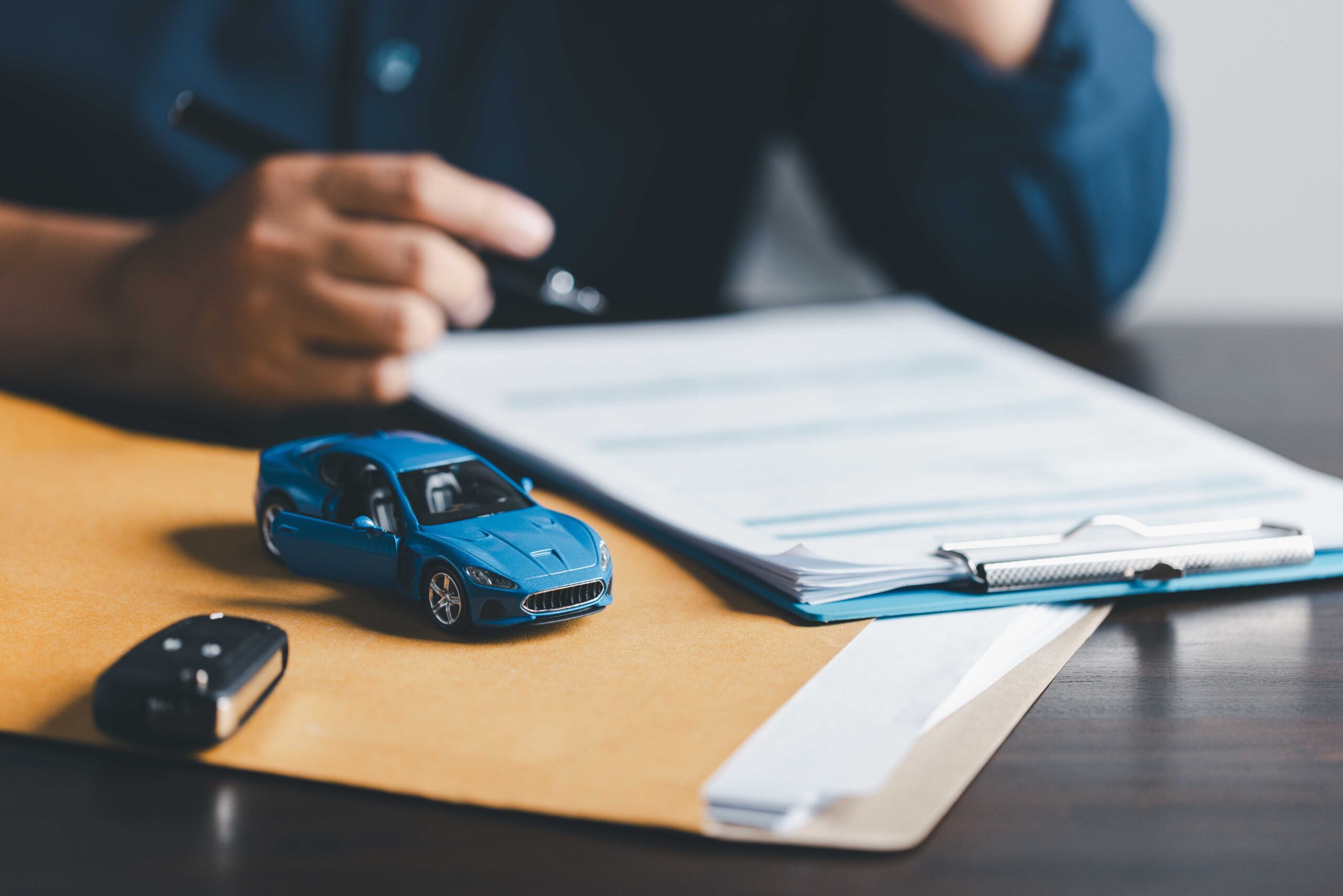 Person signing a car hire agreement document, after purchasing car rental excess insurance, attached to clipboard with blue toy car in forefront.