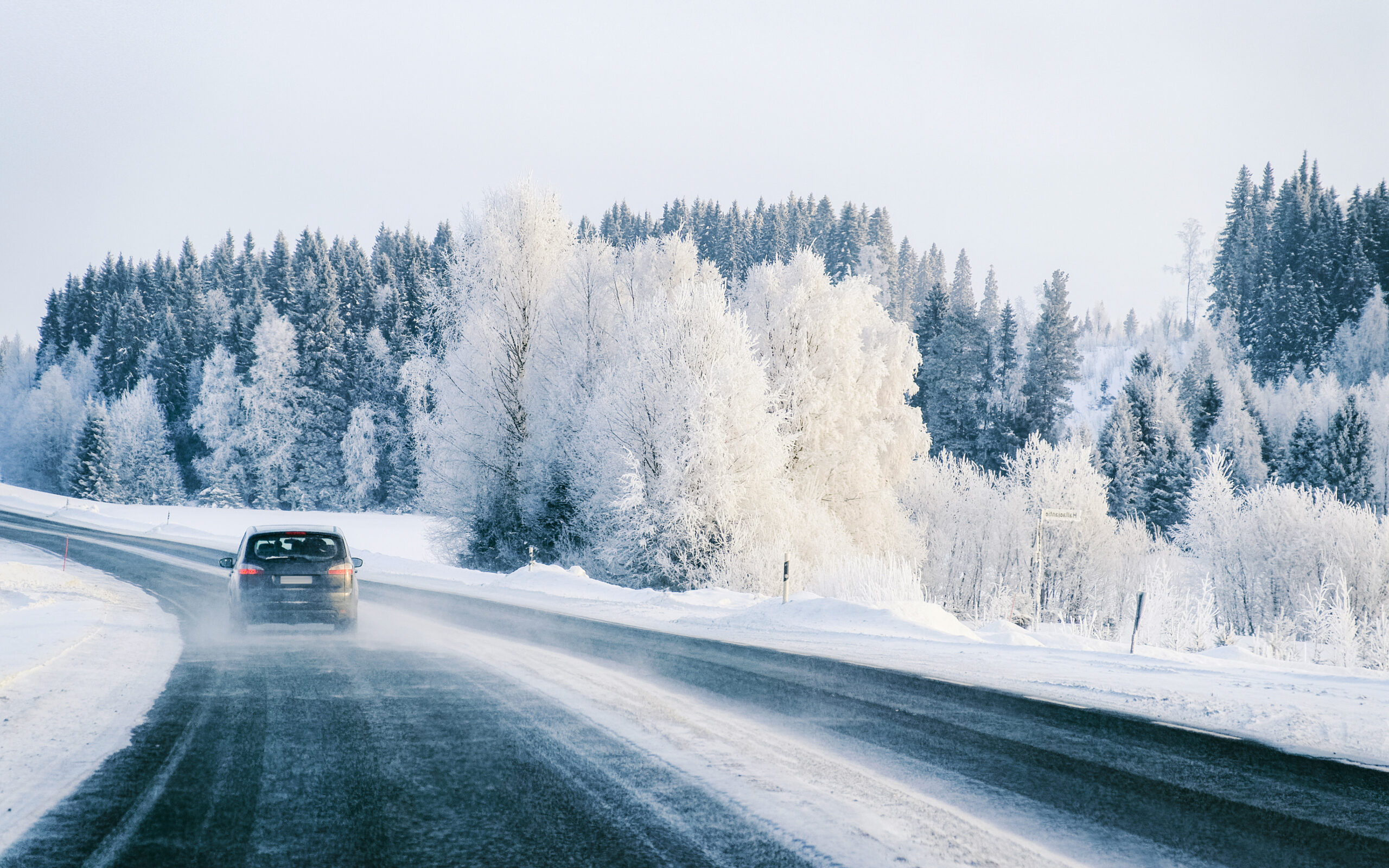 A car drives on a snowy road lined with frosted trees under a pale sky.