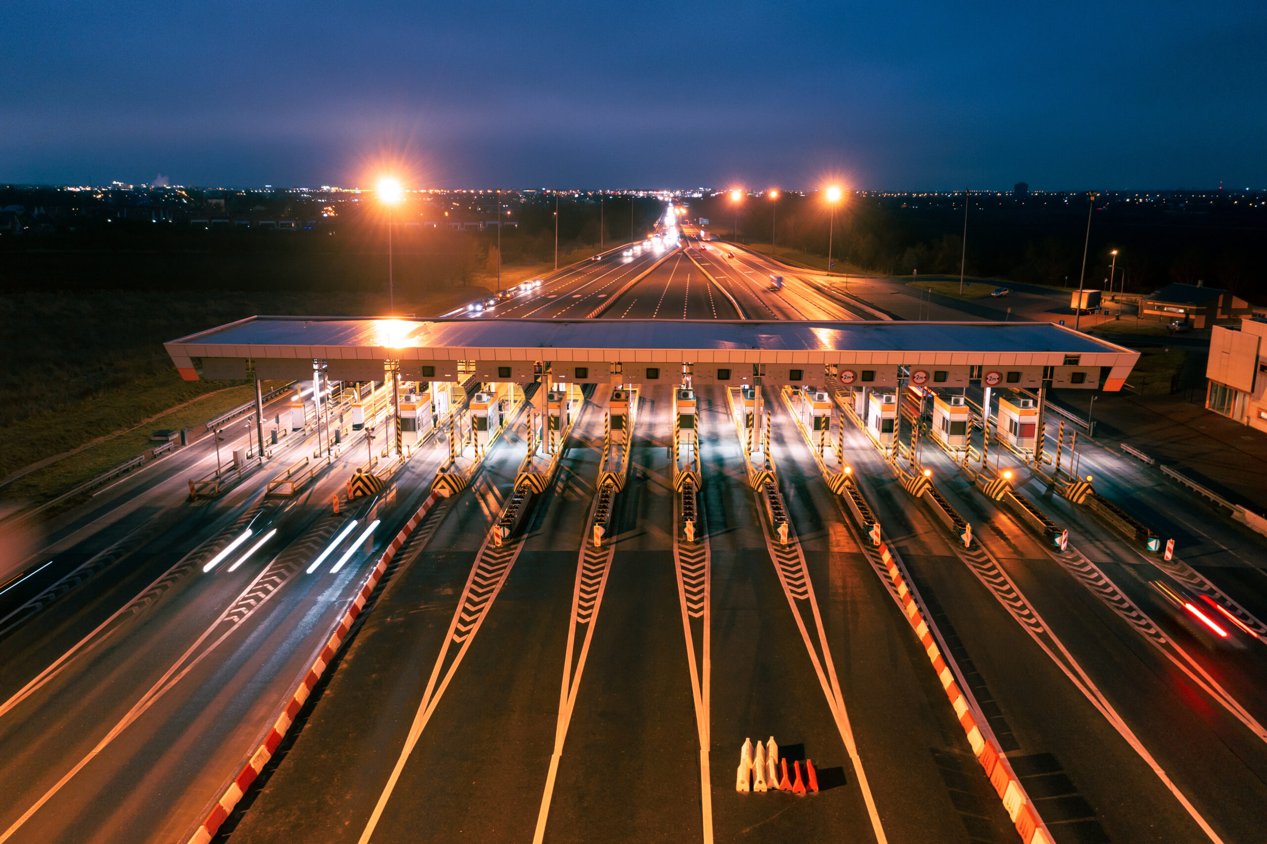 Aerial view of a brightly lit toll plaza at night with multiple lanes and blurred car lights suggesting movement.