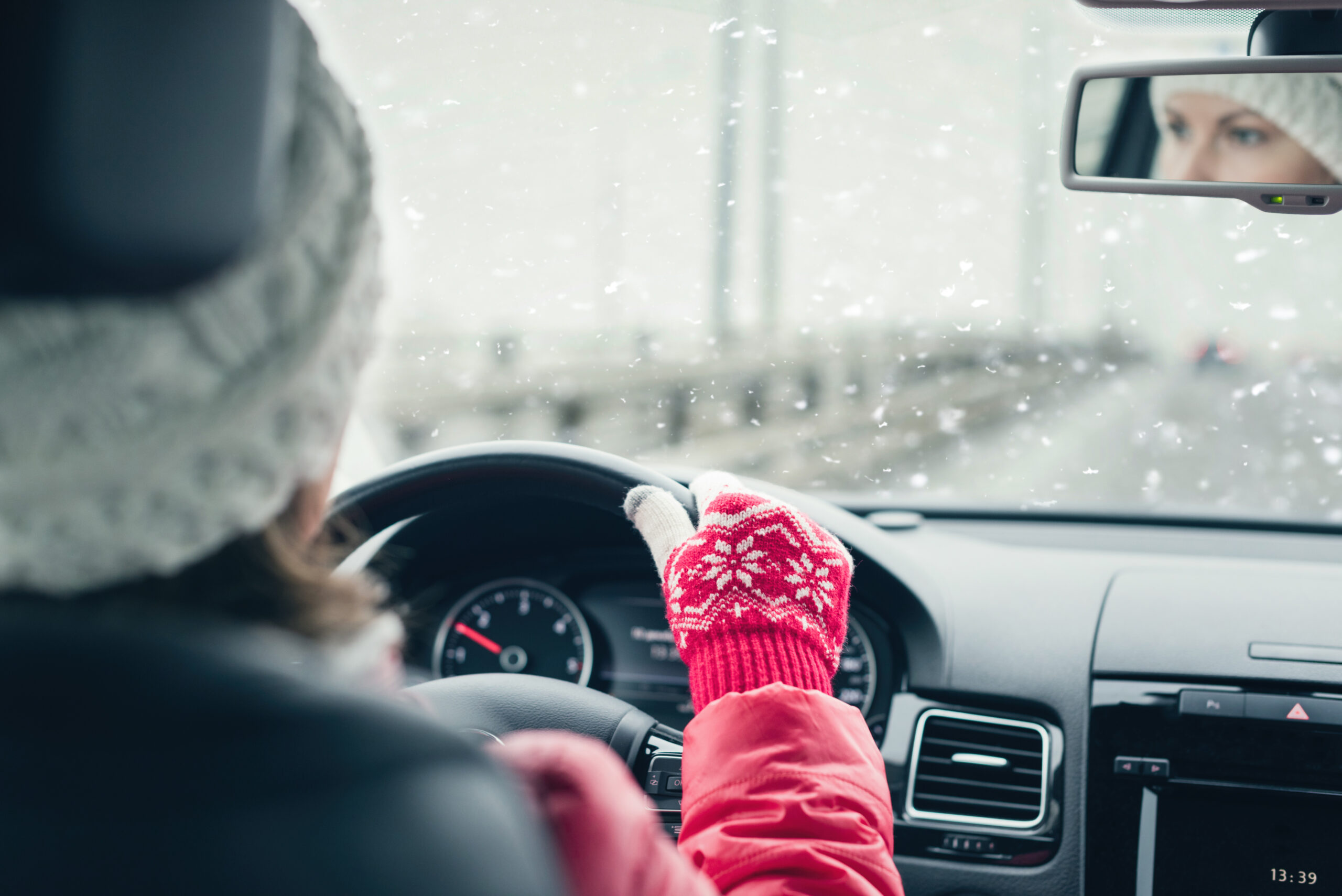 A person wearing a winter hat and red mittens drives a car on a snowy day with snowflakes visible through the windshield.
